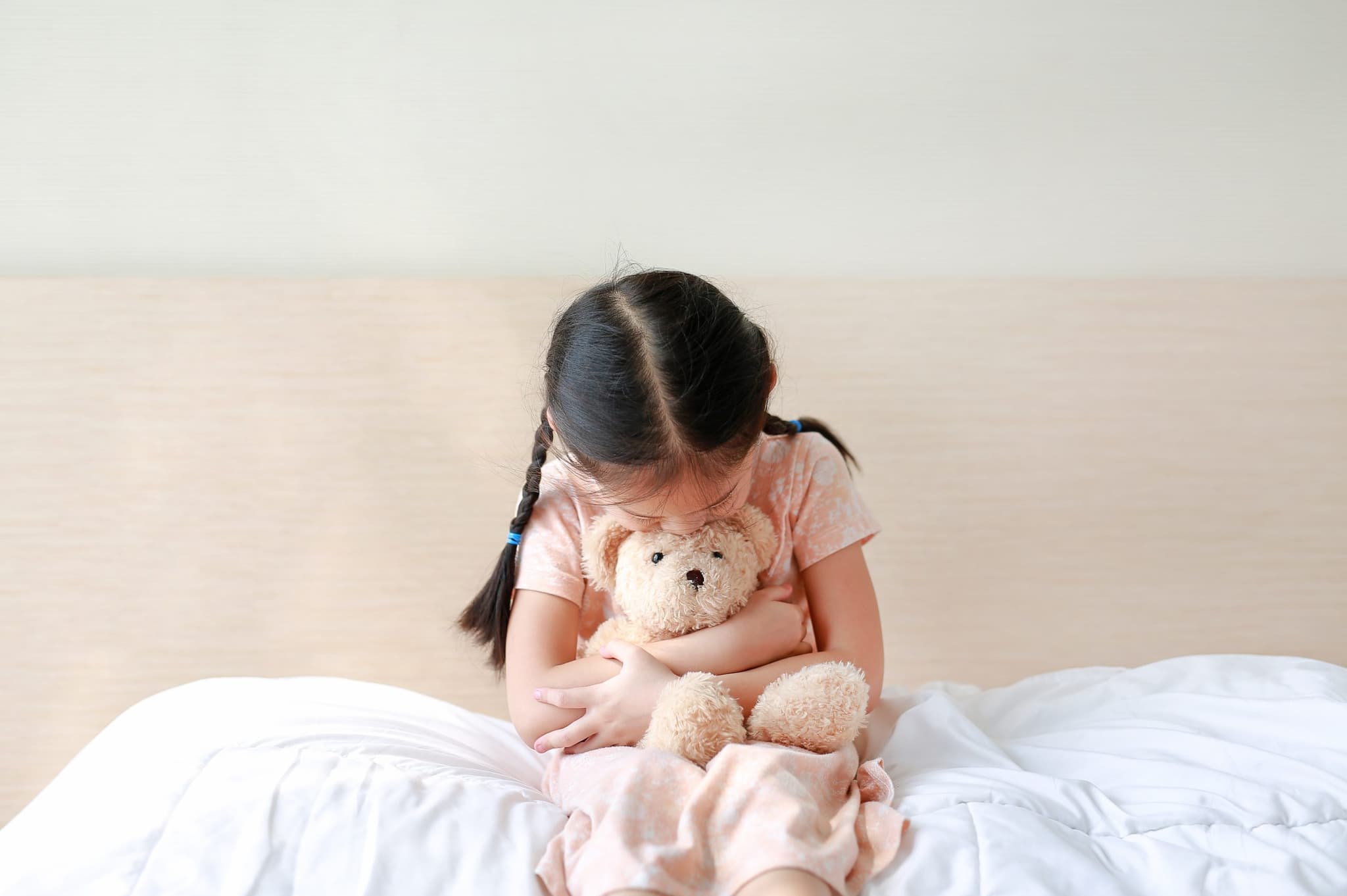 Peaceful little girl embracing teddy bear while sitting on the bed at home.