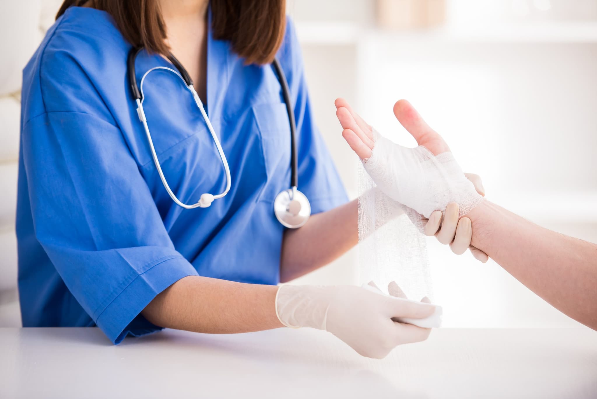 A healthcare professional in blue scrubs and latex gloves bandaging a patient's injured hand with gauze, providing medical care in a clinical setting
